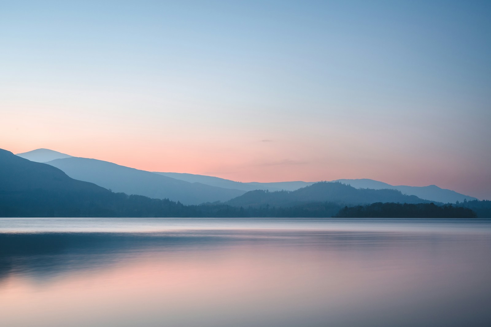 Peach sunrise reflected in a still lake beneath layers of blue mountains, photographed by Thomas Tucker